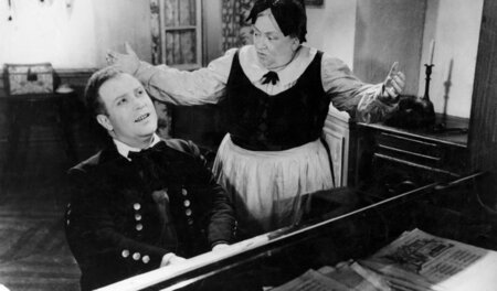 Fritz Kobus (Lucien Duboscq) am Klavier, neben ihm die Bedienste...