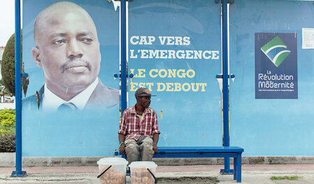Ein Straßenverkäufer sitzt in Kinshasa vor einem Portrait von Jo... Ein Straßenverkäufer sitzt in Kinshasa vor einem Portrait von Jo...